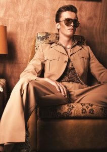 Das Bild zeigt einen sitzenden Mann mit zurückgestylter Retro-Frisur und Sonnenbrille.