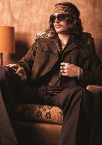 Das Bild zeigt einen sitzenden Mann mit längeren, welligen Retro-Hairstyle und Stirnband.