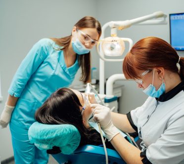 Auf dem Bild sieht man eine Patientin auf einem Zahnarztstuhl. Zwei Arzthelferinnen arbeiten an ihr.
