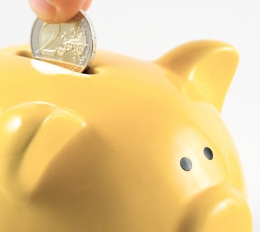 Das Bild zeigt ein gelbes Sparschwein, in das eine Hand 2 Euro steckt