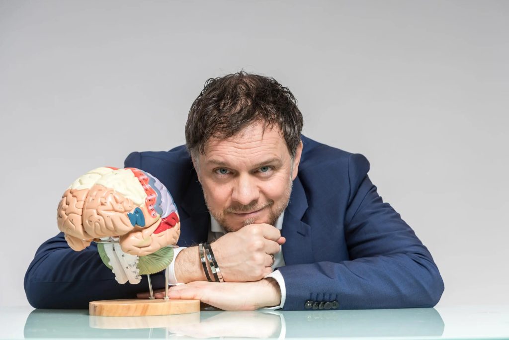 Bild von Professor Doktor Volker Busch. Die Arme liegen angewinkelt auf einem Tisch und der Kopf liegt auf den Händen. Neben ihm steht das plastische Modell eines menschlichen Gehirns