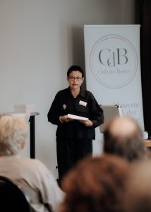 Rebecca Kandler steht vor Publikum, hat Karten in der Hand, im Hintergrund ein Banner vom Club der Besten