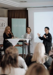 Ute Berg, Sabrina Poser und Rebecca Kandler stehen vor Publikum an einem hohen quadratischen Tisch mit weißer Tischdecke und 2 Gläsern darauf. Im Hintergrund Top Hair und Club der Besten Banner sowie eine Leinwand