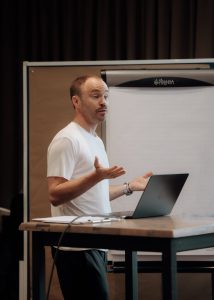 Thomas Eusemann steht an einem Pult auf dem ein Laptop und ein Klemmbrett liegt, hinter ihm steht ein Flipchart.