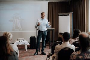 Stefan Brühl steht vor Publikum, im Hintergrund zu sehen: kleiner Tisch mit Beamer, Leinwand, Rednerpult und Flipchart