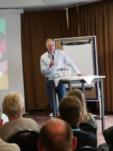 Markus Lorscheidt steht vor Publikum mit Mikrofon in der Hand an einem Rednerpult auf dem ein Laptop steht. Im Hintergrund eine Leinwand und ein Flipchart