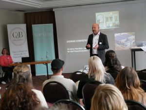 Alexander Thurner steht vor Publikum, hat Karten in der Hand und spricht. Im Hintergrund Top Hair und Club der Besten Banner, ein Tisch an dem Anke Krespach sitz und ein Laptop und eine Wasserflasche drauf steht. Zudem eine Leinwand mit Bildern der Firma Gieseke.