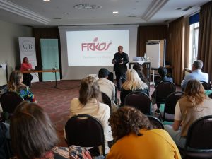 Ennio Barattucci steht vor Publikum Im Hintergrund eine Leinwand auf der "Frikos" zu lesen ist, ein Rednerpult auf dem ein Laptop, eine Flasche und ein Glas stehen. Daneben ein Top Hair und ein Club der Besten Banner, Anke Krespach sitzt an einem Tisch