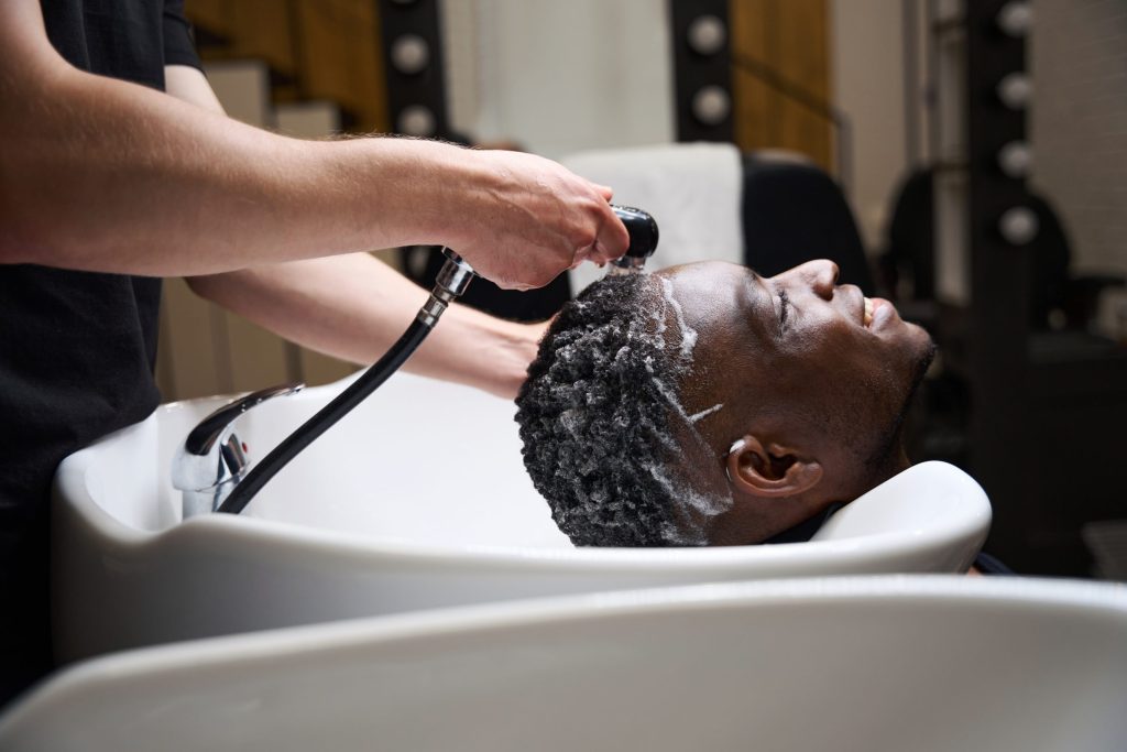 Der Mann wäscht den Schaum mit warmem Wasser vom Haar des Kunden ab, der afroamerikanische Mann hat lockiges Haar