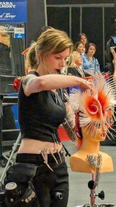 Das Bild zeigt eine Frau, die konzentriert an einem Übungskopf arbeitet. An ihrem Hosenbund stecken Haarspangen, ihren Kamm hält sie mit dem Mund fest.