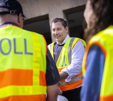 Bundesfinanzminister Lars Klingbei mit einer gelben Weste auf der Zoll steht mit zwei Zollbeamt*inen, die man von hinten sieht
