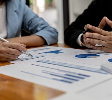 2 Menschen, von denen man nur die Hände sieht, haben ausgedruckte Tabellen und Schaubilder vor sich liegen