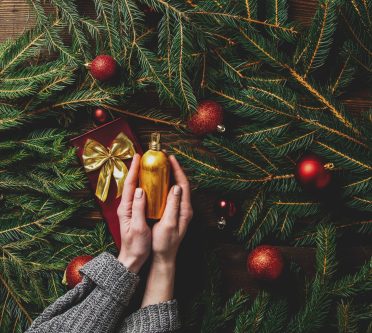 Das Bild zeigt Tannenzweige mit Christbaumkugeln und Hände, die ein Kosmetikprodukt halten