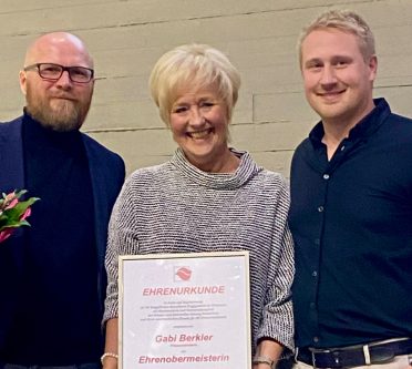 Frau in der Mitte mit Urkunde, links Mann mit Blumenstrauß, rechts noch ein Mann