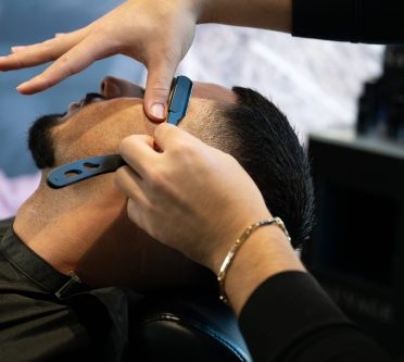 Das Bild zeigt die Hände einer Friseurin, die einem Kunden im Friseurstuhl mit einem Rasiermesser die Haarkontur erarbeitet.