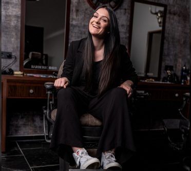 Das Foto zeigt Barberin Vanessa Hermann auf einem Barber-Stuhl in ihrem Salon sitzend und lächeln.