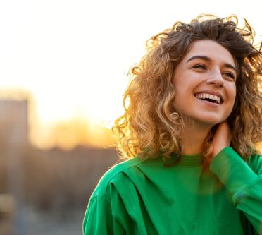 Das Bild zeigt den Kopf einer jungen Frau mit dunkelblonden Naturlocken, die lächelt, mit der linkenHand in die Haare greift und ein grünes Oberteil trägt.