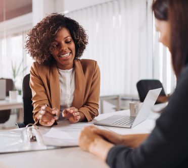 Frau sitzt einer anderen Frau gegenüber im Gespräch. Unterlagen auf dem Tisch