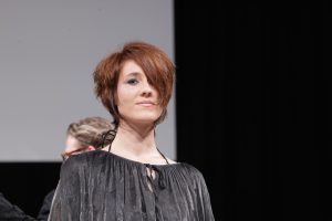 Workshop Stage 2026 27 Eine Frau mit Kurzhaarschnitt in Kupferbraun auf einer Bühne