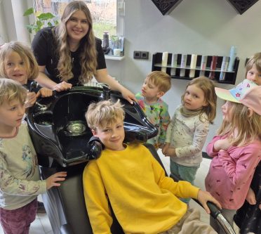 Kindergartenkinder und eine Frau stehen am Waschbecken in einem Salon. Ein Kind sitzt am Waschbecken.