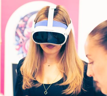 Der Blick der Kamera geht über die Schultern zweier junger Frauen zu einer blonden Frau, die eine VR-Brille trägt.