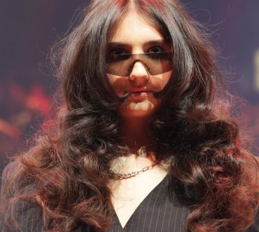 Eine Frau mit braunen, langen Haaren, die in große, glamouröse Wellen gelegt sind, trägt eine Sonnenbrille.