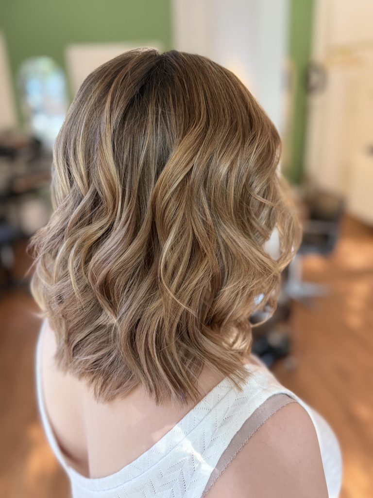 Aus der Perspektive von hinten, schräg oben werden gewellte, blonde Haare in der Farbnuance Caramel Blond gezeigt.
