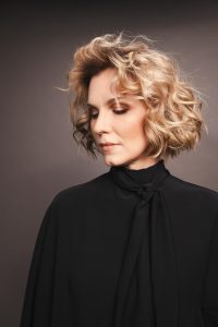 Trend Intercoiffure Austria 7 Kinnlanger, voluminöser Curly Bob in warmem Blond mit dunklerem Ansatz und lebendig definierten Locken.