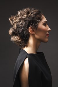 Trend Intercoiffure Austria 3 Dunkles, lockiges Haar zu einem voluminösen, unregelmäßigen Updo im Nacken zusammen gesteckt, mit helleren Akzenten in den Locken.